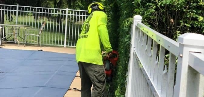 B. Woody's crew member trimming a tall hedge row along a residential fence with a hedge trimmer