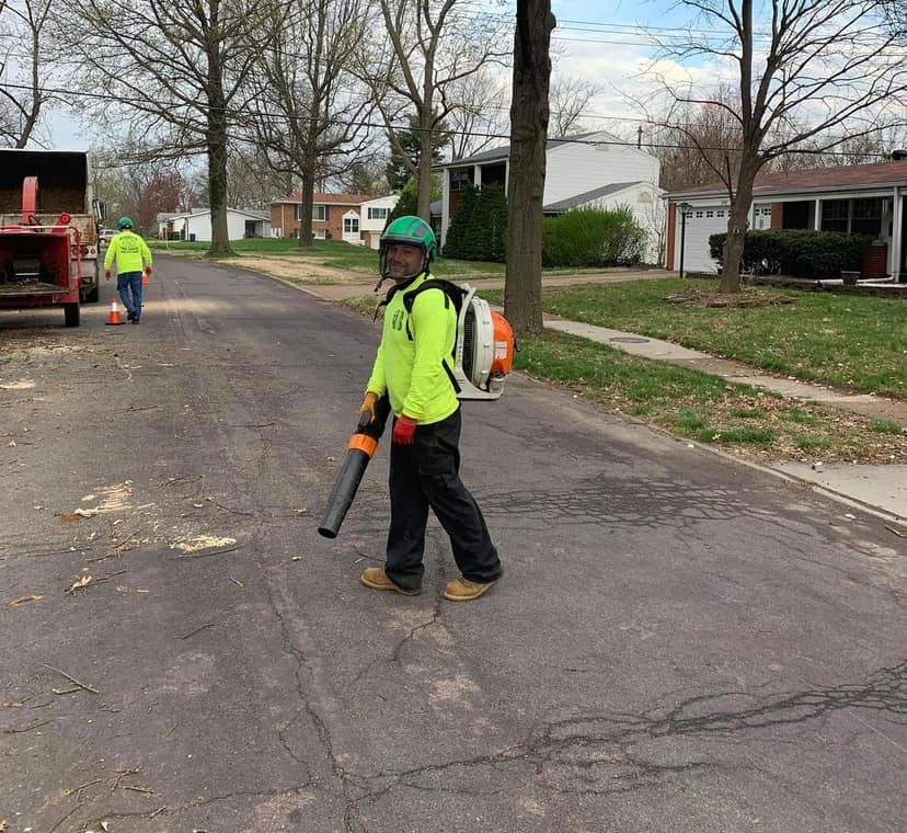 B. Woody's crew member in hi-vis gear clearing debris with a backpack blower, chipper truck and second crew member up the street